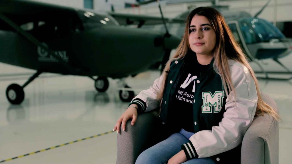 Young Woman with Long Hair Sits on a Chair in a Hangar, Wearing a Varsity-style Jacket with Patches, in Front of a Helicopter. - Los Industriales