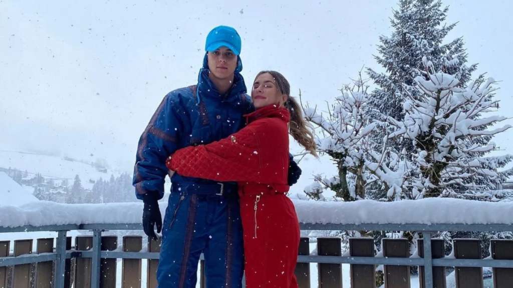 Couple in Blue and Red Winter Suits Embracing on a Snowy Balcony with Snow-covered Trees in the Background. - Los Industriales