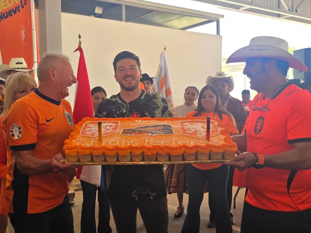 Two Men in Orange Team Shirts Hold a Large Orange Celebration Cake As Others Cheer in a Crowded Event Space. - Los Industriales